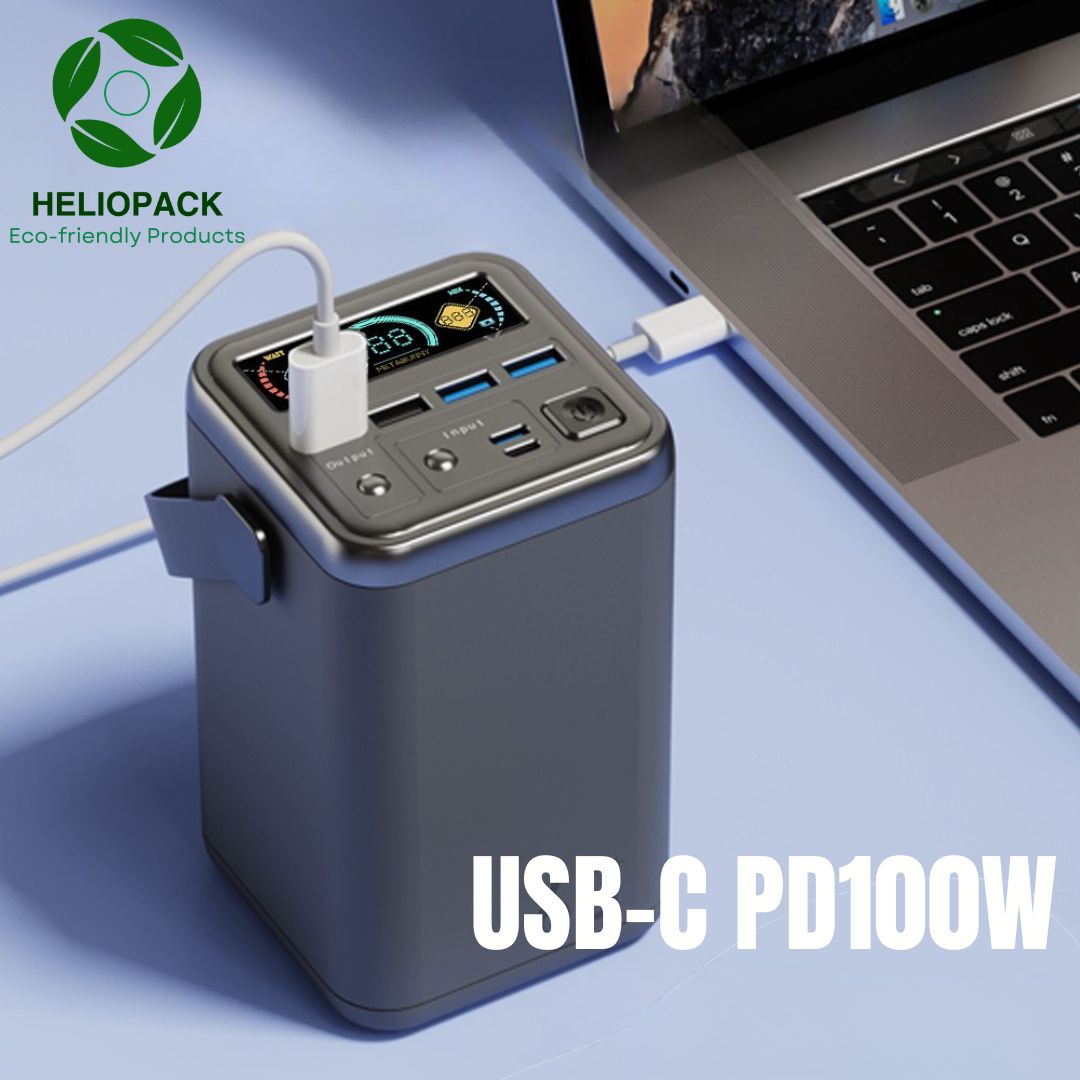 Image publicitaire de la batterie externe haute capacité HELIOPACK 60000mAh PD100W, connectée via un câble USB-C à un ordinateur portable ouvert sur un bureau. Un écran d'affichage numérique actif, des ports multiples et le texte "USB-C PD100W" en bas.